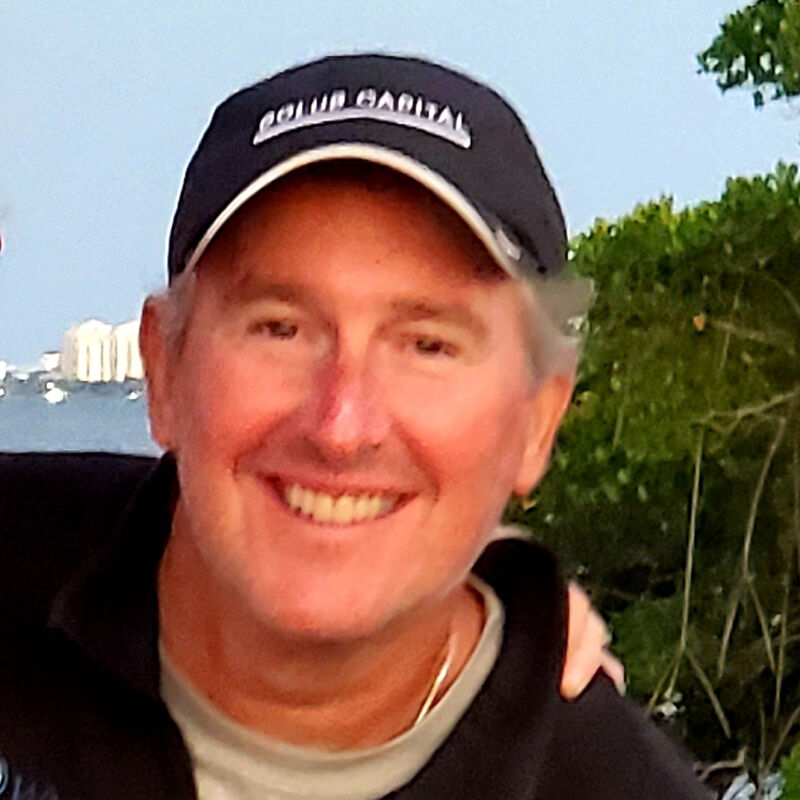 The image shows a middle-aged man with fair skin, wearing a dark baseball cap with white lettering and a white brim. He is smiling broadly, revealing his teeth. The background is slightly blurred, suggesting an outdoor setting, possibly near water, with some greenery visible.